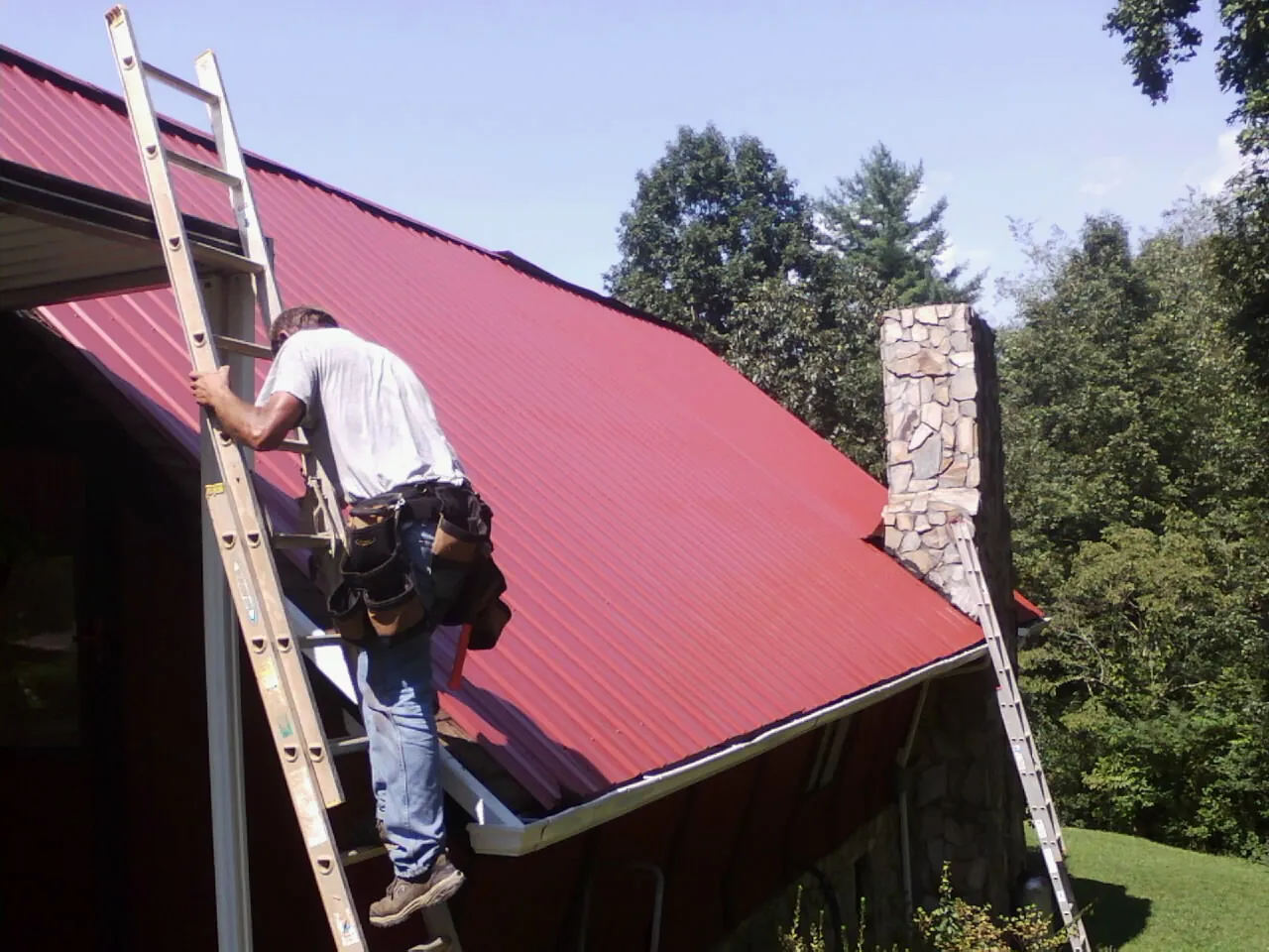 Roofer on a ladder beside a red metal roof and stone chimney — Soffit & Fascia Repair by Roofing Brothers in Denver